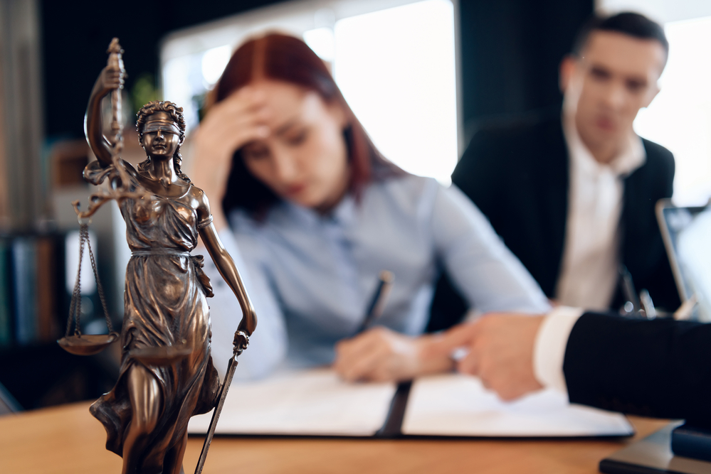 bronze statue of themis holds scales of justice. in unfocused background, couple signs documents.